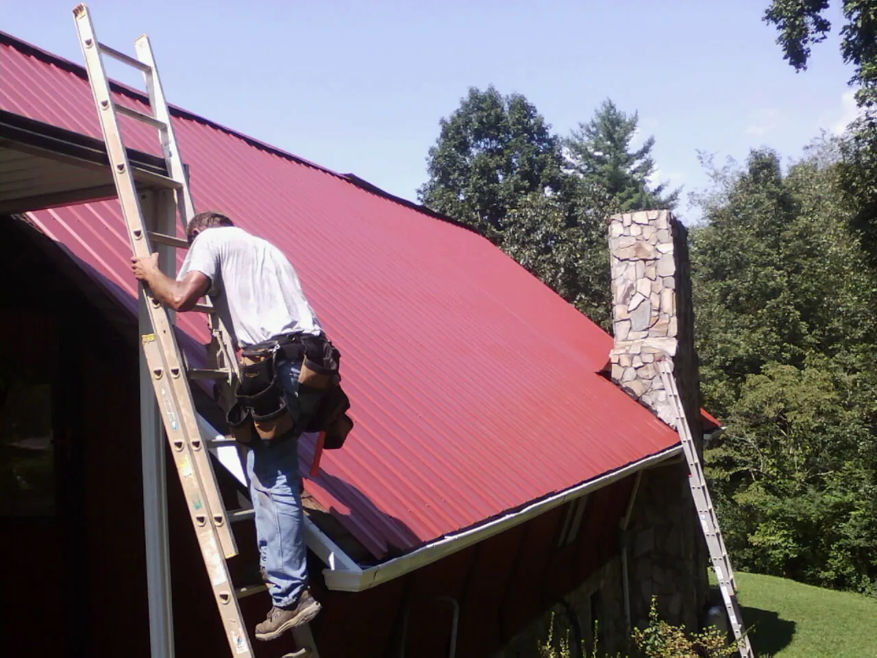 Roofer on a ladder beside a red metal roof and stone chimney — Soffit & Fascia Repair by Roofing Brothers in Dittmer